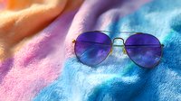Stylish aviator sunglasses with purple gradient lenses on colorful tie-dye textile background combining pink, orange and blue colors in soft dreamy summer mood.
