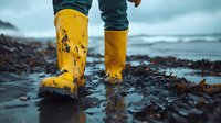 Person wearing bright yellow rubber boots walking on wet rocky beach shore with ocean waves in background during overcast weather conditions.