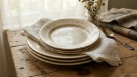 Stack of cream ceramic plates with linen napkin on rustic wooden table near window, styled for dining and tableware design projects.