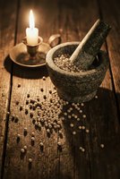 Stone mortar and pestle with scattered peppercorns on rustic wooden table beside flickering candle creating warm atmospheric cooking scene.