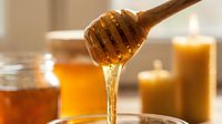 Golden honey dripping from wooden dipper into glass bowl with jars and candles creating warm amber lighting for natural organic food photography.