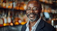 Mature African American businessman with glasses smiling in upscale bar with blurred bottles background, dressed in elegant blazer and white shirt.
