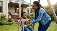African American mother pushing happy toddler son on wooden swing in sunny backyard garden. Family bonding time outdoors at home with laughter and joy.
