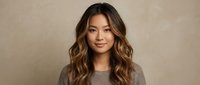 Young Asian woman with long wavy brown hair wearing casual sweater against neutral beige background for lifestyle and portrait photography.