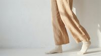 Person walking in beige linen wide-leg pants and white socks against a bright white wall, casual minimalist fashion lifestyle concept.