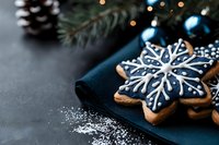Festive snowflake-shaped gingerbread cookie decorated with navy blue icing and white details on dark background with pine branches and Christmas lights bokeh.