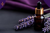 Amber glass dropper bottle with essential oil next to dried lavender sprigs on dark surface with purple silk fabric background for aromatherapy and wellness.