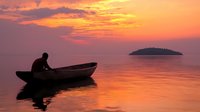 Silhouette of person in wooden boat during dramatic orange sunset over calm water with small island on horizon, reflecting golden light on tranquil sea surface.