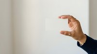 Man's hand holding blank white card against neutral wall background for business messaging and branding purposes.