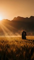 Romantic couple silhouette embracing in golden wheat field at sunset with dramatic sunbeams and mountain backdrop for wedding and love concepts.