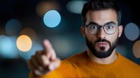 Middle Eastern man with beard and glasses showing pointing gesture against blurred bokeh background, wearing orange sweater. Surprised facial expression.