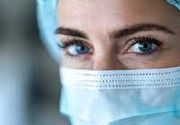 Close up portrait of medical professional wearing surgical mask and cap, with intense blue eyes and freckles, looking directly at camera in hospital setting.