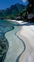 Pristine tropical beach with crystal clear turquoise water, white sand, palm trees and traditional thatched huts against dramatic mountain backdrop for travel content.