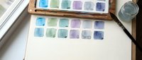 Watercolor paint palette with blue green purple color swatches on white paper near window. Art supplies setup for creative painting and color mixing projects.