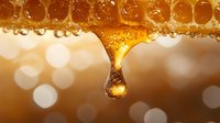 Golden honey dripping from honeycomb with natural bokeh background, macro close-up showing viscous drop formation and crystalline texture in warm amber tones.