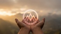 Hands holding glowing sphere with mountain logo against dramatic sunset sky and mountain landscape for branding and corporate identity concepts.