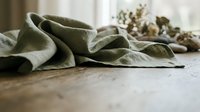 Soft olive green linen napkins arranged on rustic wooden table with dried eucalyptus branches creating warm natural dining atmosphere for home decor styling.
