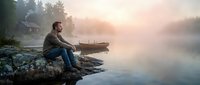 Man sitting on rocky shore beside wooden boat at misty lake during golden hour sunrise creating peaceful outdoor recreation scene.