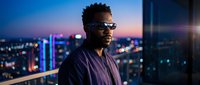 Young Black male professional wearing sunglasses against illuminated city skyline at dusk. Urban nightlife backdrop with bokeh lights. Corporate, tech, and lifestyle use.