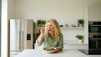 Young blonde woman enjoying healthy meal in modern white kitchen with green plants and stainless steel appliances during daytime.