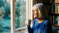 Senior woman with gray bob haircut enjoying morning coffee by window in cozy home library with bookshelves, peaceful retirement lifestyle.