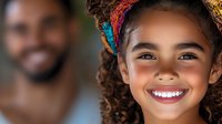 Young mixed-race girl with curly hair and colorful headband smiling brightly, showing white teeth against blurred background of male figure. Natural lighting portrait.