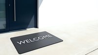 Dark welcome mat with white lettering placed on a light concrete porch in front of a modern navy blue front door.