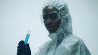Laboratory scientist in protective hazmat suit and goggles examining blue liquid in test tube against foggy background, conveying research and scientific discovery.