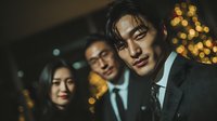 Young Asian businessman in formal suit smiling confidently at camera with colleagues in background during evening corporate event with warm bokeh lights.