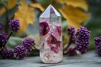 Pink crystal point with natural inclusions stands against blurred purple flowers and autumn leaves background, showcasing mystical mineral formation.