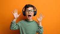 Excited young boy with glasses and headphones expressing surprise against vibrant orange background, wearing green striped shirt.
