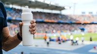 Close-up of athlete's hand holding blank white sports drink bottle at stadium during match, ideal for beverage marketing and sports hydration campaigns.