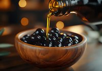 Pouring golden olive oil into wooden bowl filled with fresh black olives, creating appetizing Mediterranean scene with warm bokeh background lighting.