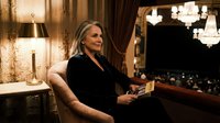 Elegant blonde woman in black dress relaxing with book in luxurious hotel suite with warm lighting and classic decor for lifestyle and travel concepts.