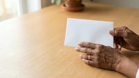 Senior adult with dark skin tone holding blank white card on wooden table surface, suitable for mockups, business communications, and greeting card designs.