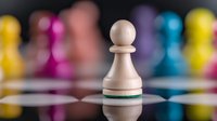 White wooden pawn chess piece with green stripe stands prominently on reflective surface against blurred colorful chess pieces background for strategy games.