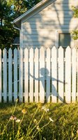 White picket fence casting shadow on light blue house exterior with wildflowers in foreground, residential property and suburban home exterior design element.