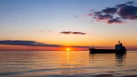 Cargo ship silhouette on calm ocean waters during golden sunset with dramatic sky and sun reflection creating peaceful maritime scene.