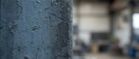 Weathered blue paint peeling off concrete pillar in industrial warehouse setting with blurred background machinery and equipment.