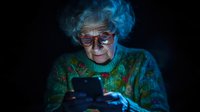 Elderly Caucasian woman with blue hair using smartphone in dark room with glowing blue light illuminating her face and colorful knitted sweater.
