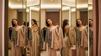Young women with diverse ethnicities trying on neutral-toned dresses and blazers in a modern retail fitting room with multiple mirrors, shopping and fashion retail concept.