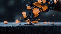 Dramatic macro shot of orange fragments shattering against dark textured surface, creating dynamic contrast between warm and cool tones in moody atmosphere.