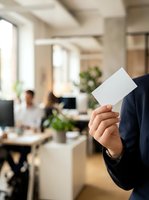 Businessman in dark suit holding blank white business card in modern office environment with colleagues working at desks and green plants.