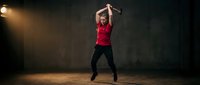 Athletic young woman jumping with arms raised in red tank top and black leggings during fitness workout in dramatic gym lighting for sports and exercise concepts.