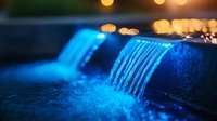 Illuminated waterfall with glowing blue water cascading over dark stone edge at night, creating magical atmosphere with bokeh lights and reflections.