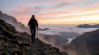 Hiker with trekking poles standing on mountain slope at sunrise above clouds, scenic alpine landscape with misty valleys below.