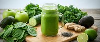 Fresh green smoothie in a mason jar surrounded by spinach, kale, avocado, apple, cucumber, lime, ginger, and chia seeds on a wooden board.