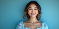 Young Asian woman with bright smile wearing light blue lace dress against turquoise background, natural makeup and wavy hairstyle expressing joy and confidence.