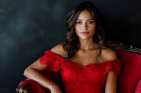 Young Hispanic woman in red lace off-shoulder dress sitting elegantly on vintage velvet armchair against dark dramatic background, soft studio lighting highlights natural makeup.