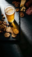 Golden beer glass with gourmet cheese platter, salami, nuts and pretzels on dark slate board for appetizer menu and pub dining experience.